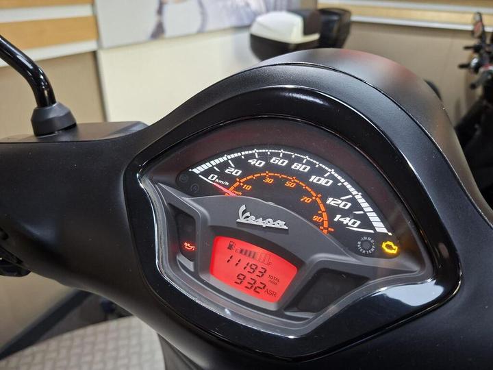 Close-up of a Vespa scooter's dashboard, showing a speedometer with a maximum speed of 140 km/h, mileage of 1193 miles, and a digital display. Warning lights are visible, including those for maintenance and traction control.