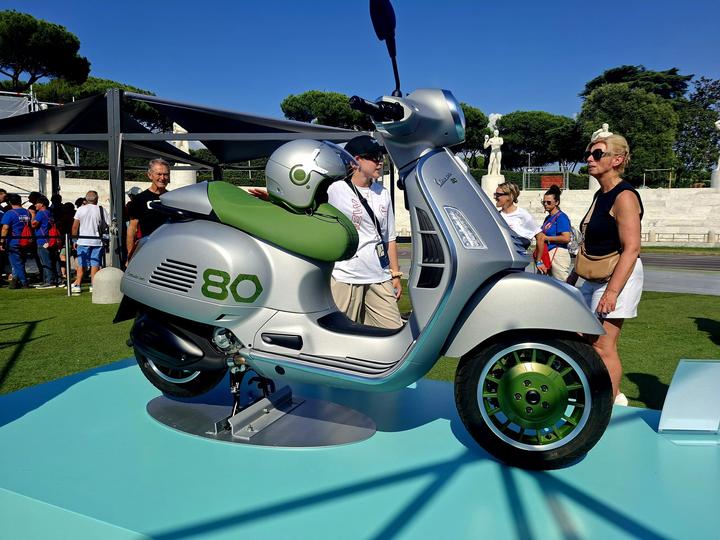 A silver vintage-style Vespa scooter with a green seat and matching wheels is displayed on a turquoise pedestal, surrounded by people in a sunny outdoor setting.