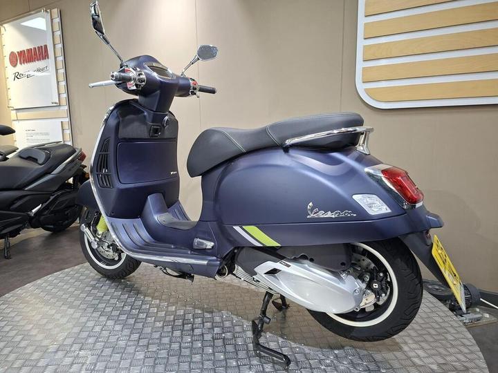 A blue Vespa scooter displayed in a showroom, showcasing its sleek design and chrome details, with a textured floor and neutral wall background.