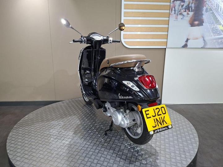 A sleek black Vespa scooter with a brown seat is displayed on a circular metallic platform, showcasing its rear side and license plate reading "EJ20 JNK." The backdrop features a minimalist interior design.