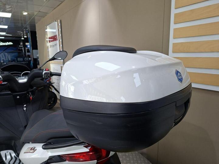 A close-up view of a white motorcycle top case mounted on the rear of a scooter, with part of the scooter's seat and handlebars visible in the foreground, set against a blurred showroom background.