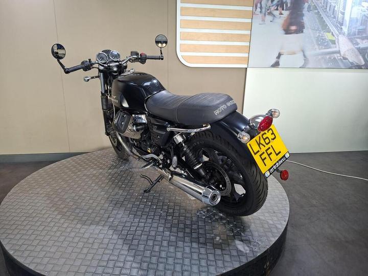 A black Moto Guzzi motorcycle displayed on a circular metal platform, viewed from the rear angle, showing its sleek design, distinctive logo on the seat, and rear license plate. The background features a light-colored wall with vertical stripes and a digital display of people in an urban setting.