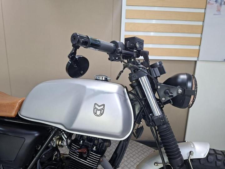 A close-up view of a motorcycle's handlebars, featuring a matte silver fuel tank, a brown leather seat, a black headlight, and side mirrors. The motorcycle has a minimalist design with visible cables and controls.