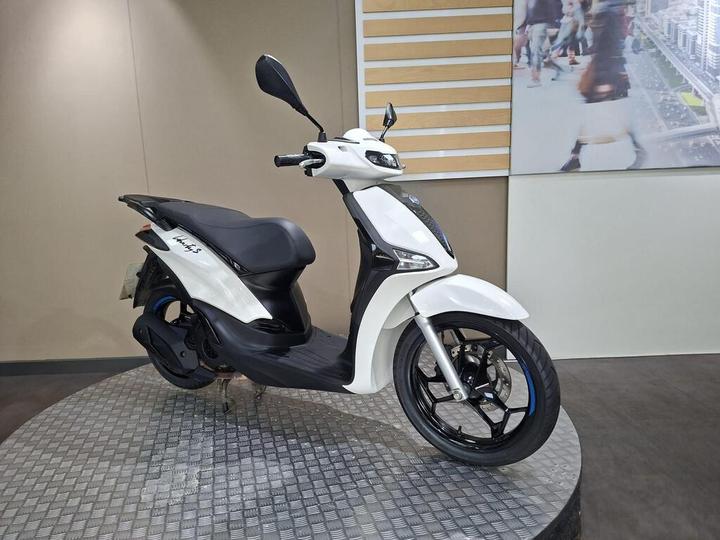 A white scooter with a sleek design is displayed on a circular metallic platform, set against a plain backdrop with a modern image of a busy urban scene.
