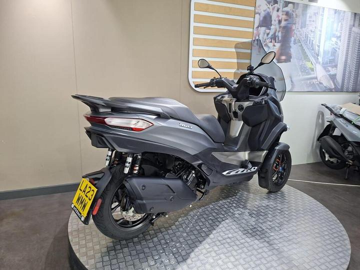 A black and gray three-wheeled scooter displayed on a circular platform, with a blurred background showing a cityscape. The scooter features a sleek design, highlighted by its headlights and rear tail lights. A license plate at the back reads "L23 WMH."