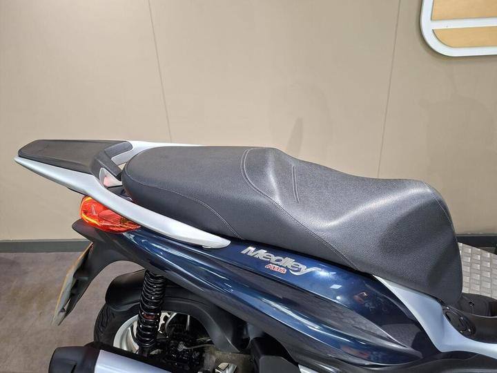 Close-up of a motorcycle seat on a scooter with a dark leather texture, featuring a silver trim and red taillight, against a neutral background.