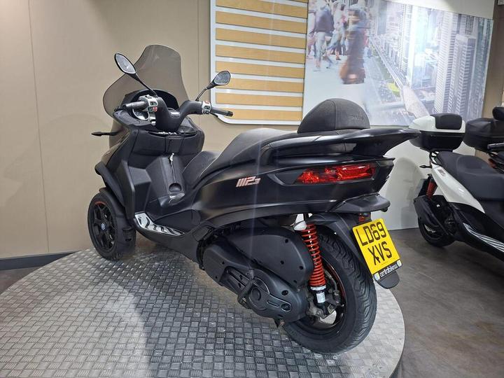 A black three-wheeled scooter with a clear windshield, parked on a patterned floor in a showroom. The scooter features red suspension springs and has a visible license plate labeled "LD69 XVS." In the background, a city scene mural is displayed.