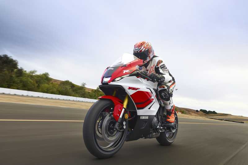 A motorcyclist in a racing suit and helmet rides a red and white Yamaha YZF-R motorcycle on a paved road, surrounded by greenery and a cloudy sky.