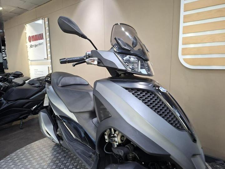 A sleek, modern scooter with a dark gray body and clear windshield is displayed in a showroom, with other scooters visible in the background and a Yamaha sign on the wall.