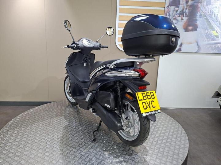 A black scooter with a blue storage case on the rear, displayed on a circular platform, against a neutral background. The license plate is visible, reading "LB68 OVC."