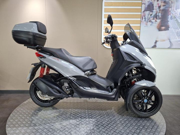 Piaggio MP3 300HPE Sport (2020) A sleek gray three-wheeled scooter with a black storage case on the rear, featuring a modern design and red suspension springs, displayed on a circular patterned platform against a neutral background.