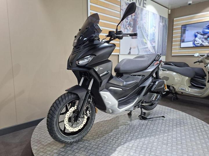 A black and silver scooter with a prominent windscreen is displayed on a textured platform in a modern showroom, with a light-colored scooter visible in the background.