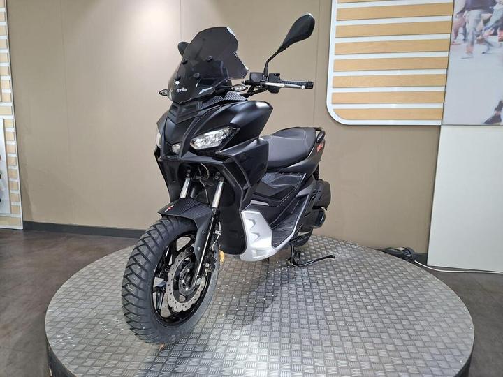 A sleek black scooter displayed on a rotating platform, featuring off-road tires, a large windshield, and modern design elements against a neutral background.