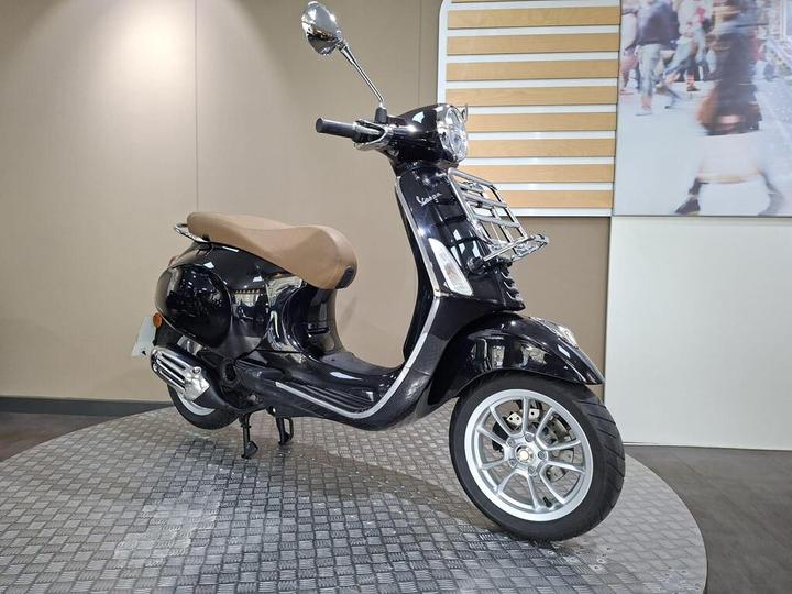 A sleek black Vespa scooter with a tan seat, displayed on a circular, textured platform against a neutral backdrop.