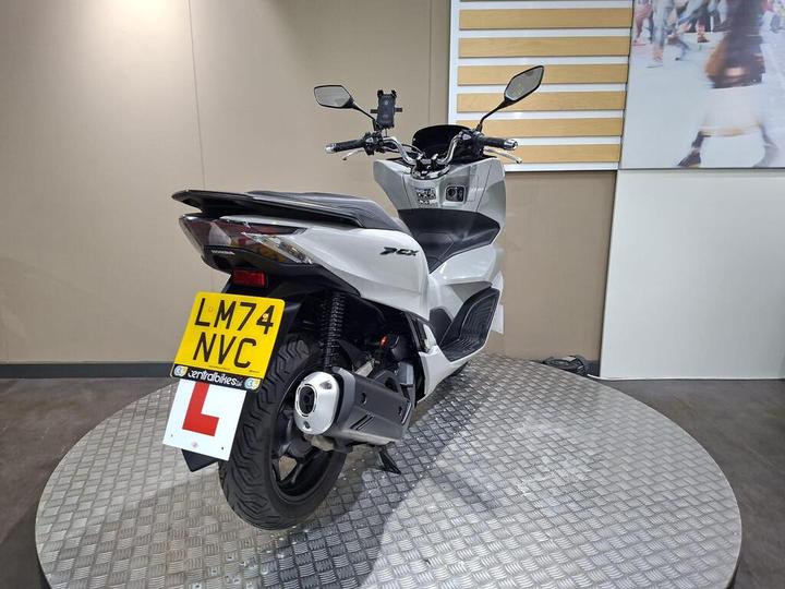 A white Honda PCX motorcycle displayed on a circular metal platform, with a visible license plate reading "LM74 NVC" and a learner driver sign. The background features a plain wall and a larger image of people in motion.