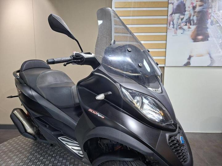 A black scooter with a windshield is displayed in a showroom, showcasing its sleek design and modern features. The background includes a blurred image of people walking.