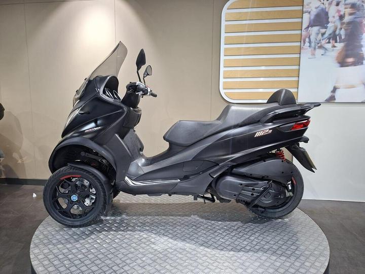 A black three-wheeled scooter displayed on a circular platform with a modern interior backdrop.
