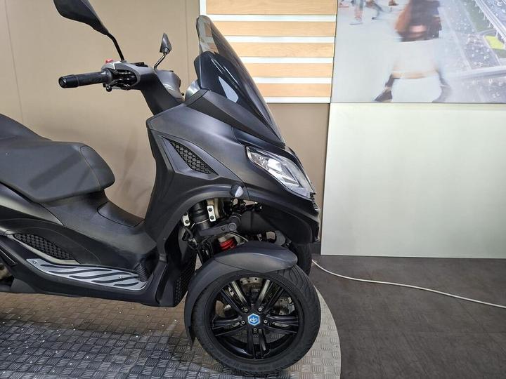 A sleek black scooter or three-wheeled motorcycle parked on a textured flooring, with a modern backdrop of a wall featuring a wooden panel and a digital display.