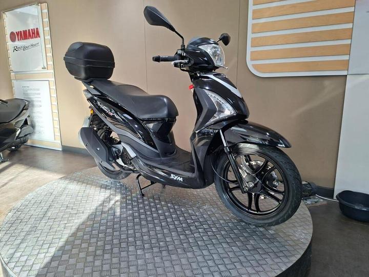 A black SYM scooter is displayed on a circular metallic platform inside a showroom, with a Yamaha sign visible in the background. The scooter features a top case and a sleek design.