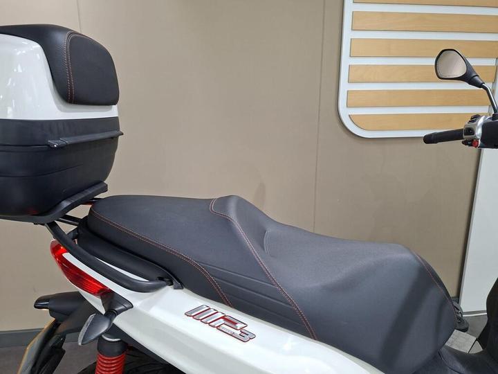 A close-up view of a white scooter featuring a black seat with red stitching and a rear storage box. The background is neutral with wooden paneling.