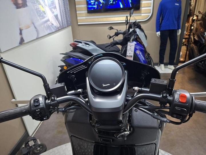 A close-up view of a motorcycle's handlebars and dashboard, featuring a circular speedometer, control switches on both sides, and a background displaying other motorcycles and a mannequin in a blue shirt.