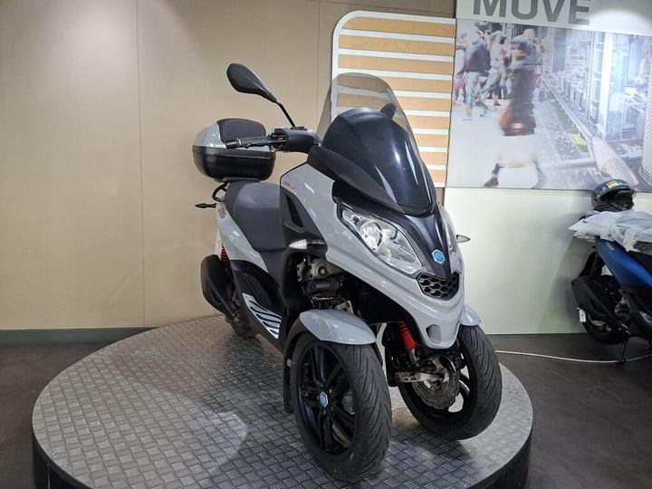 A gray and black three-wheeled scooter displayed on a circular stand in a showroom, with a sleek design and modern features, set against a neutral wall background.