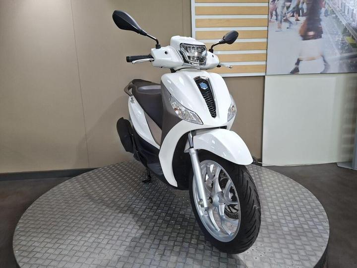 A white scooter displayed on a circular platform, featuring a sleek design with a prominent front headlight and modern mirrors, against a neutral backdrop.