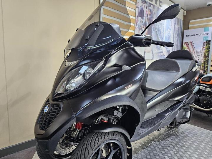 A sleek black scooter with a streamlined design, featuring a tinted windshield, prominent headlights, and a sporty appearance, displayed in a showroom setting.