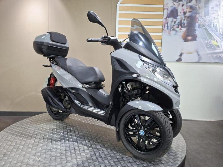 A modern three-wheeled scooter in gray, featuring a black seat, side panel accents, and a top case for storage, displayed on a textured circular platform against a neutral background.