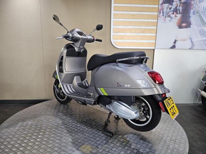 A gray scooter is displayed on a circular platform with a textured surface, featuring a retro design, chrome mirrors, and a license plate reading "V173 CET." The background includes a light-colored wall and a blurred image of people and roadways.