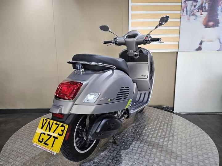 A sleek gray scooter with a green accent, displayed on a circular metal platform, showcasing its rear and side view, with the license plate visible. The background features a neutral wall and an abstract image.