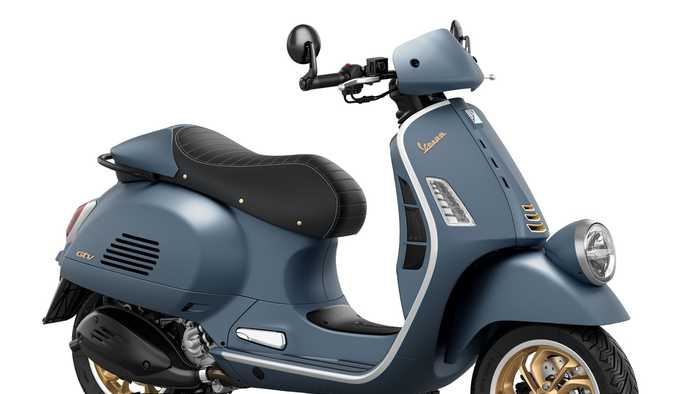 A blue Vespa scooter with a black seat, gold rims, and stylish chrome details, positioned at a slight angle to showcase its classic design.