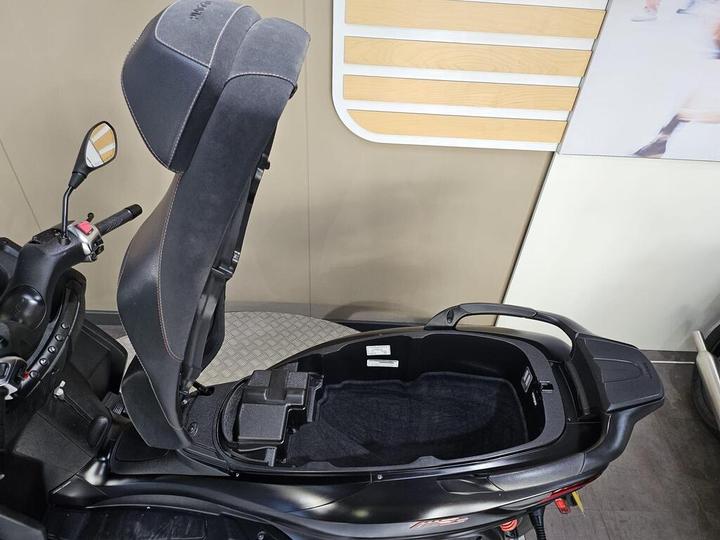 A close-up view of an open scooter seat, revealing a spacious storage compartment underneath. The seat is upholstered in black fabric with stitching details, and the scooter's dashboard and controls are visible on the left. A wall with linear design patterns is in the background.