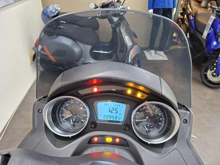 Dashboard of a Piaggio scooter displaying speedometer, tachometer, and digital readout with odometer information. In the background, a scooter and a mannequin wearing a blue shirt are visible.