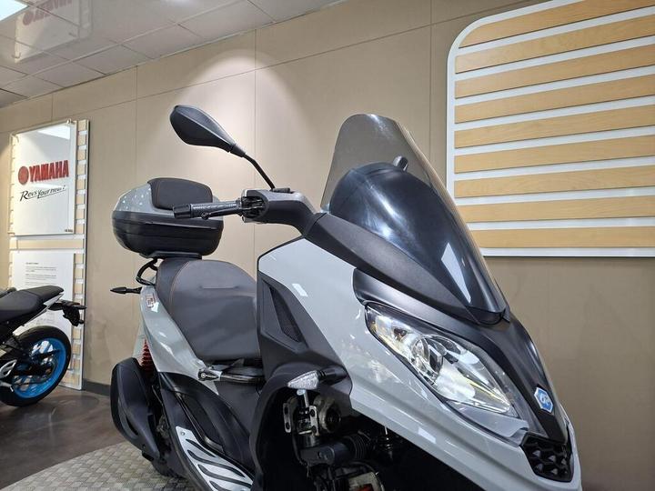 A close-up view of a gray and black Yamaha scooter, featuring a large windshield, side mirrors, and a rear storage compartment, displayed in a showroom with promotional Yamaha signage in the background.