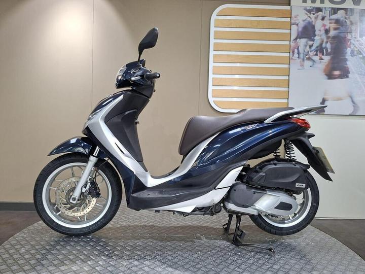 A blue and white scooter is displayed in profile on a textured surface, with a neutral wall in the background featuring a design element.