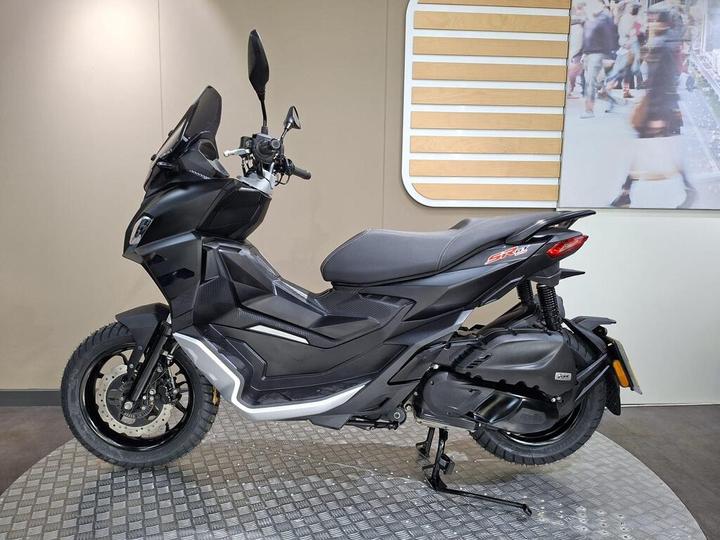 A sleek black scooter displayed in profile, featuring a modern design with a prominent front, two wheels, and a side stand, set against a neutral background.