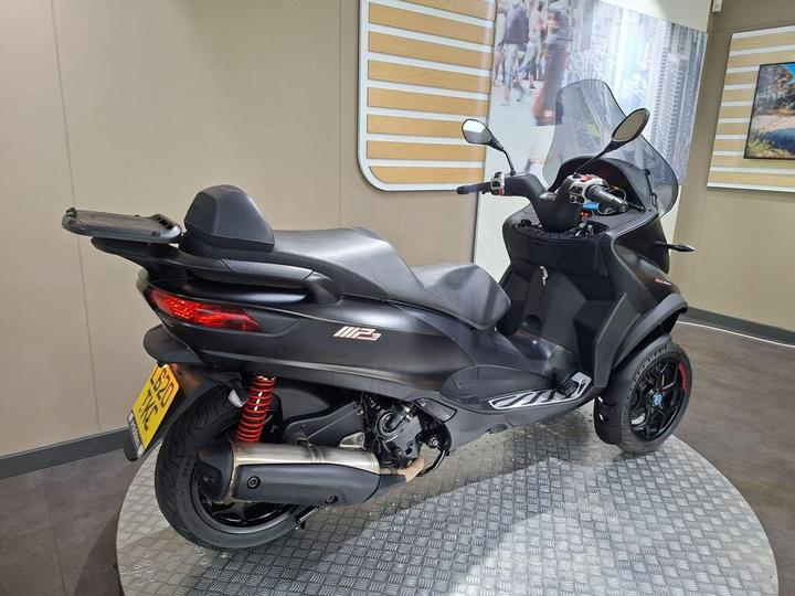 A sleek black three-wheeled scooter with red shock absorbers, displayed in a modern interior setting.