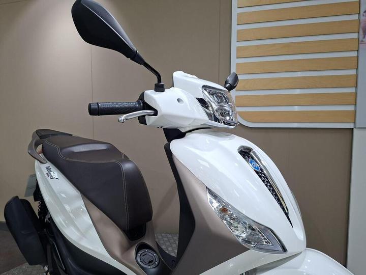 A close-up of a white scooter with a brown seat, featuring a rearview mirror on the left side and a sleek headlight design. The background is plain and minimalistic, emphasizing the scooter's design elements.
