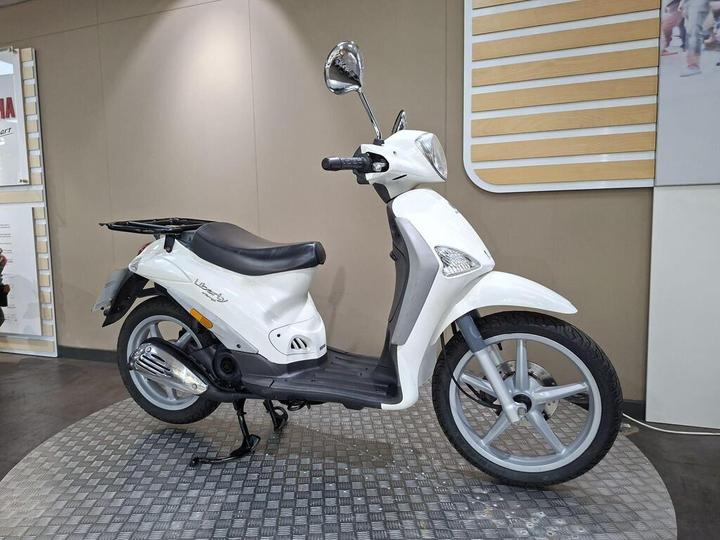 A white scooter displayed on a textured circular platform, featuring a black seat, mirror, and modern design elements, set against a neutral background.