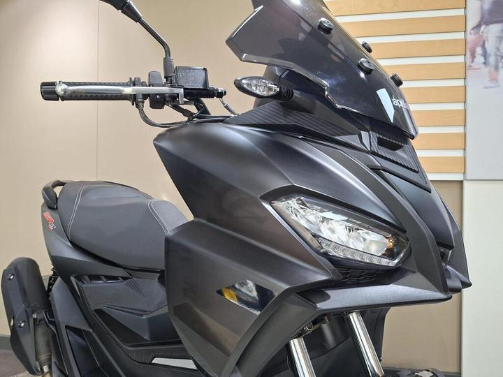 A close-up view of a modern black scooter showcasing its sleek design, including the handlebar, front fairing, headlights, and windscreen. The background features a neutral-colored wall with a wooden panel.