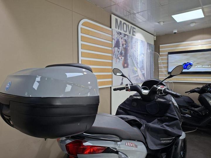 A close-up view of a gray scooter with a large rear storage box, positioned in a modern display area featuring a "MOVE" sign and a monitor showing motorcycle racing footage.