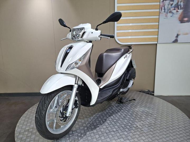 A white scooter is displayed on a circular, textured platform, featuring a sleek design with a brown seat and chrome accents, against a neutral background.