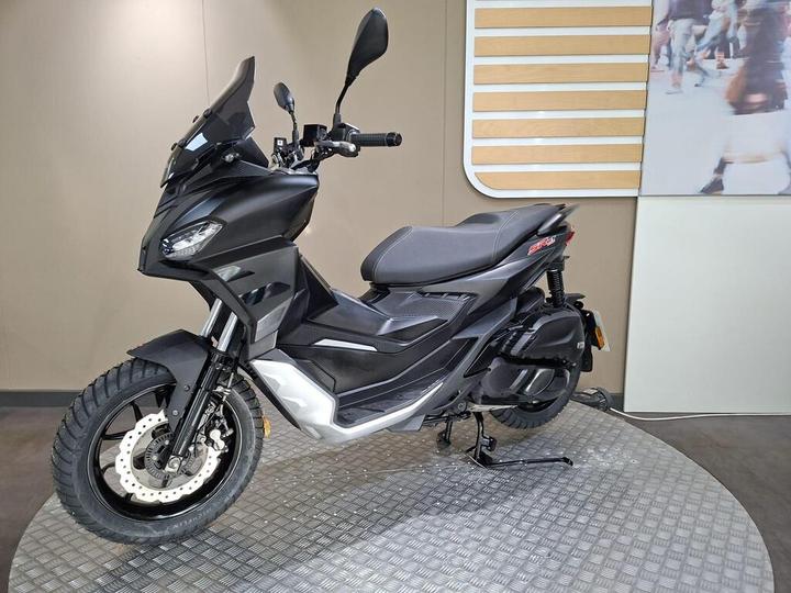 A sleek black scooter with a tall windshield and rugged tires, showcased on a circular platform in a modern setting, featuring a light background.