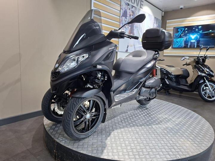 A black three-wheeled scooter displayed on a circular platform inside a showroom, with a large windshield and storage compartment on the back. The background features light-colored walls and other scooters visible.