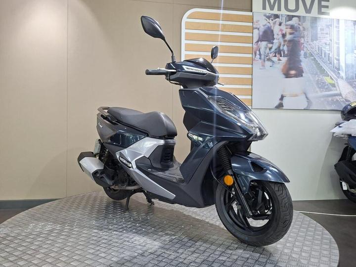A sleek dark blue scooter parked on a textured platform, featuring a modern design, front mirror, and visible branding. The background includes a light-colored wall and a blurred image of people, suggesting a lively urban environment.