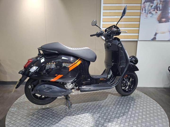 A black scooter with orange stripe accents is displayed on a circular metal platform in a modern showroom. The scooter is viewed from the side, showcasing its sleek design and glossy finish.