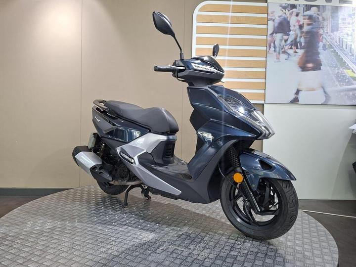 A modern scooter in dark blue and silver, displayed on a circular metal platform, with a neutral background featuring wooden accents and an image of a bustling street scene.