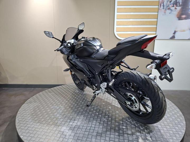 A sleek black motorcycle is displayed on a circular platform with a textured surface. The bike is shown from a rear three-quarter angle, highlighting its sporty design, curved tail, and large rear tire. The background features a neutral-colored wall with a stylized image in the corner.