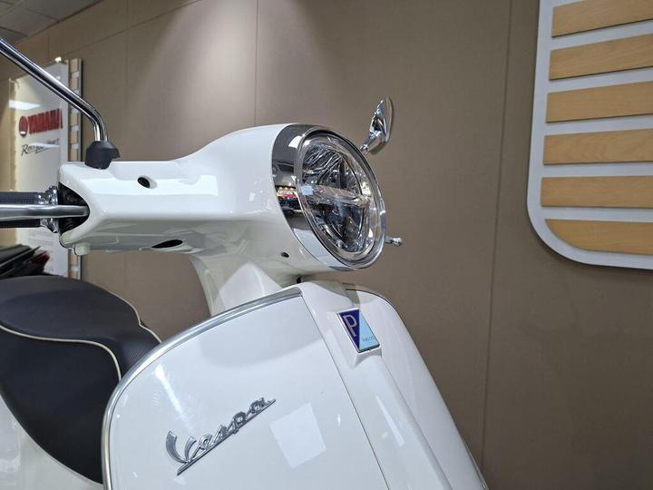 Close-up of a white Vespa scooter's front, showcasing the round headlight, handlebar, and the glossy body with the Vespa logo. A blue parking sticker is visible on the front panel against a neutral background.